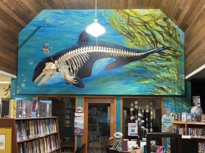 a mural with a painting of a whale behind a skeleton of a whale in a library 