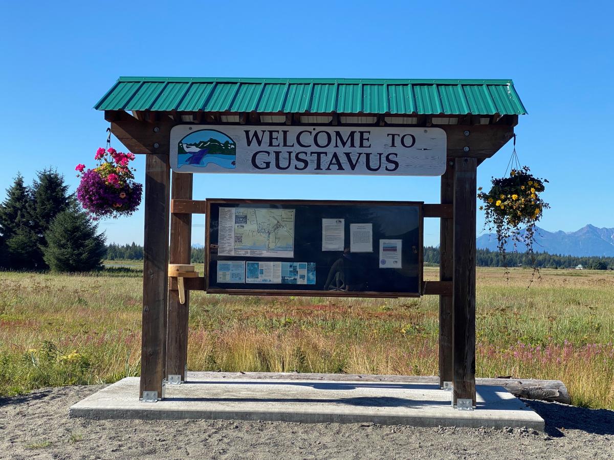 Photo of message board that says welcome to Gustavus with mountains in the background 
